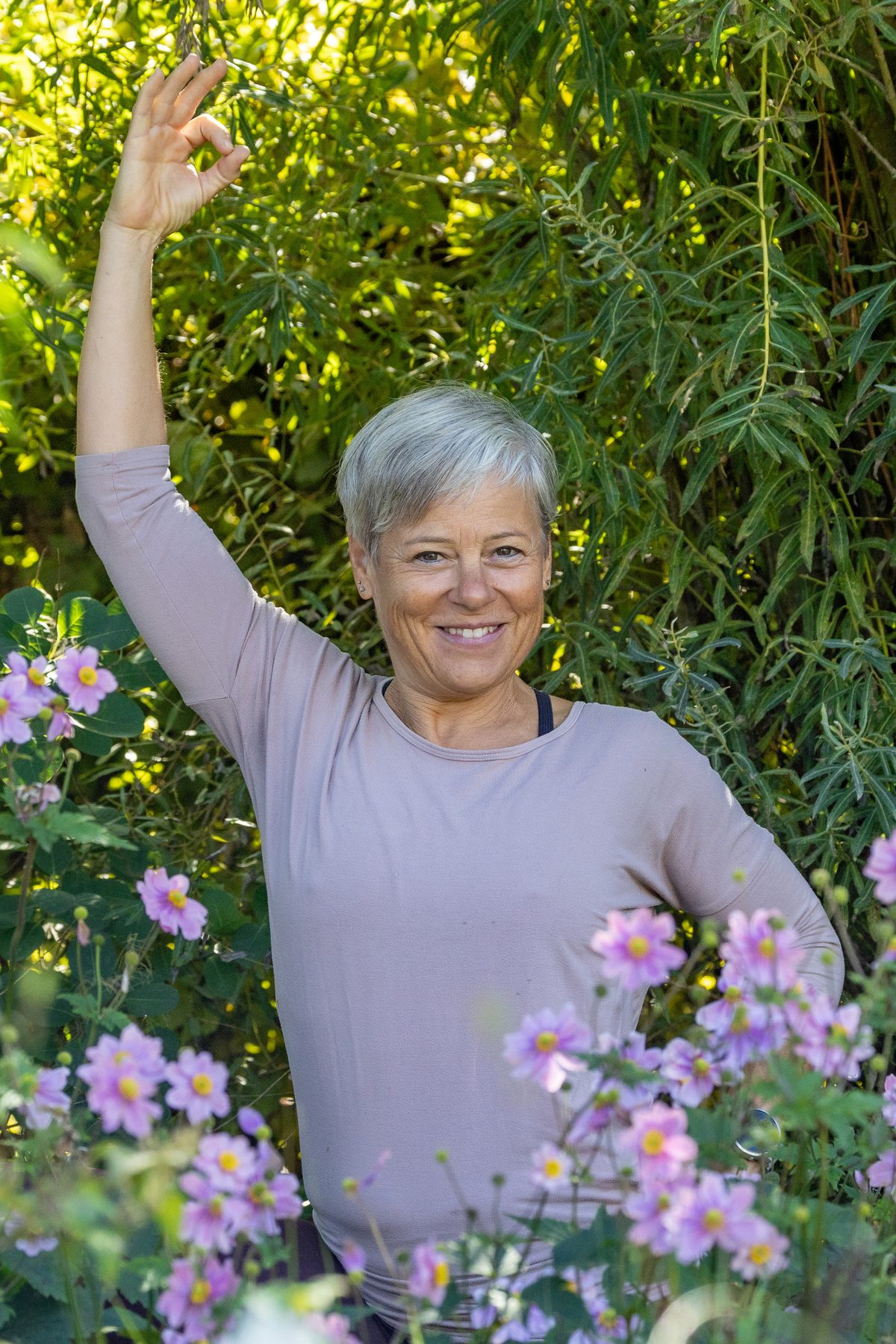 Astrid Michelle Leu mit erhobenem Arm und Mudra, lächelnd zwischen rosa Anemonen