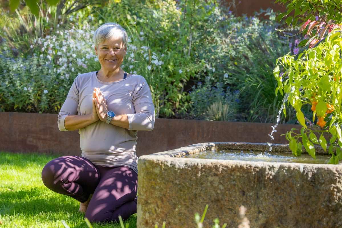 Astrid Michelle Leu sitzt mit gefalteten Händen an einem Brunnen im Garten